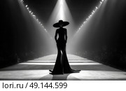 Silhouetted fashion model in dramatic spotlight walking runway in black haute couture gown and wide-brim hat during stylish monochrome fashion show. Стоковое AI-изображение, AI-художник Катерина Бондарец / Фотобанк Лори