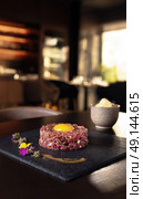 Gourmet beef tartare with raw egg yolk, beautifully plated with edible flowers and sauce, served on black slate in upscale restaurant setting. Стоковое AI-изображение, AI-художник Катерина Бондарец / Фотобанк Лори