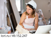 Young girl in beige dress and protective helmet compares plan of renovated premises with project on the laptop screen. Стоковое фото, фотограф Яков Филимонов / Фотобанк Лори