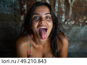 Young woman in a colorful dress playfully sticks her tongue out while indoors against a rustic, textured background. Стоковое AI-изображение, AI-художник Данила Большаков / Фотобанк Лори