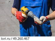 A worker in a blue jumpsuit holds an electric screwdriver with a riveter attachment and an exhaust rivet against a slate wall. Стоковое фото, фотограф Неведицына Елена / Фотобанк Лори