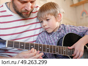Handsome dad teaches his cute son to play the guitar sitting on sofa in the living room at home. Happy Father's Day. Father teaching his child to play guitar. Kid Learning music skill. Art education. Стоковое фото, фотограф Марина Демидюк / Фотобанк Лори
