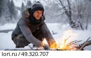 Survivalist building a fire in the open snow, wearing extreme cold weather gear. Life in permafrost conditions, climate change problems on the planet. Global warming. Стоковое AI-изображение, AI-художник ALLA YURTAYEVA / Фотобанк Лори