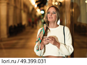 Blonde woman with smartphone in the street. Defocused city lights at the background. Pretty girl with pigtail hairstyle at night. Стоковое фото, агентство Ingram Publishing / Фотобанк Лори