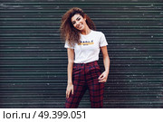 Smiling young arabic woman with black curly hairstyle. Arab girl in casual clothes in the street. Happy female wearing white t-shirt and checked pants against urban blinds. Стоковое фото, агентство Ingram Publishing / Фотобанк Лори