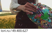 The close-up image shows a colorful palette in the hands of an artist in a stained apron as she mixes paints to paint a landscape. Стоковое видео, видеограф Strunov David / Фотобанк Лори
