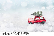 Christmas tree on roof of red toy retro car in snow through snowy winter wonder land with snowfall. Creative Christmas holiday concept. Copy space for your greeting and design. Front view. Стоковое фото, агентство Ingram Publishing / Фотобанк Лори