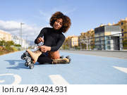 Young smiling black girl sitting on bike line and puts on skates. Woman with afro hairstyle rollerblading on sunny day. Стоковое фото, агентство Ingram Publishing / Фотобанк Лори