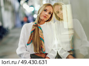 Blonde girl wearing white sweater standing in the street with defocused city lights at the background. Pretty woman with pigtail hairstyle at night reflected in a shop window. Стоковое фото, агентство Ingram Publishing / Фотобанк Лори