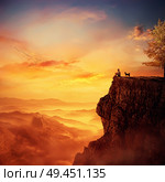 Young man with his faithful dog standing on the peak of a cliff watching the sunset over valley. Recalling childhood memories, friendship between human and animal. Стоковое фото, агентство Ingram Publishing / Фотобанк Лори
