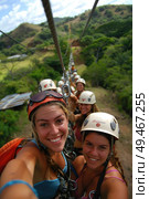 A group of individuals wearing helmets and harnesses ride along a zip line, dangling from a suspended cable high above the ground. Стоковое AI-изображение, AI-художник Данила Большаков / Фотобанк Лори
