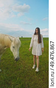 Lovely young girl is offering white horse some grass from her hand, standing in midst of a verdant meadow in the summer. Vertical. Стоковое видео, видеограф Strunov David / Фотобанк Лори