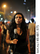 A young woman holding a microphone on a bustling street at night, with blurred passersby in the background. Стоковое AI-изображение, AI-художник Данила Большаков / Фотобанк Лори