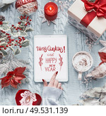 Female hand touches tablet computer with Merry Christmas and Happy New Year greeting card on light blue knitted blanket with red Christmas decoration and burning candles. Top view. Flat lay. Стоковое фото, агентство Ingram Publishing / Фотобанк Лори