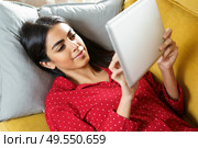Young woman wearing pyjamas at home using digital tablet on a couch. Persian woman at home using digital tablet. Стоковое фото, агентство Ingram Publishing / Фотобанк Лори