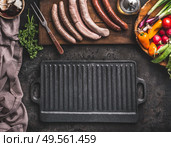 Grill or barbecue food background. Empty cast iron grill griddle and meat fork on rustic kitchen table. Various sausages on a wooden cutting board. Wooden bowl with vegetables. Top view. Стоковое фото, агентство Ingram Publishing / Фотобанк Лори