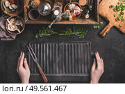 Grill or BBQ food background. Woman hold empty cast iron grill griddle and meat fork on roast rustic kitchen table. Wooden box with seasonings, oils and other kitchen utensils.Top view. Place for text. Стоковое фото, агентство Ingram Publishing / Фотобанк Лори