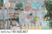 Elderly woman with gray hair holds a medicated shampoo for hair loss in hands and scans the barcode on her mobile. Pensioner pays for purchases at a pharmacy using a QR code in a mobile application. Стоковое видео, видеограф Яков Филимонов / Фотобанк Лори