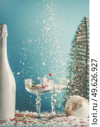 Christmas and New Year’s Eve background with champagne glasses and white champagne bottle. Christmas bauble and Christmas tree at blue background with snow and confetti. Front view. Стоковое фото, агентство Ingram Publishing / Фотобанк Лори
