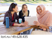 Group of positive young multiethnic female friends smiling and drinking coffee, to go while watching funny video on tablet, during break in outdoor cafe in park. Cheerful diverse ladies sharing tablet during coffee break in cafe. Стоковое фото, агентство Ingram Publishing / Фотобанк Лори