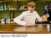 Bearded man in white shirt and cap removing napkin holder and wiping counter with cloth while working in bar in evening. Bartender cleaning counter in a nice pub. Стоковое фото, агентство Ingram Publishing / Фотобанк Лори