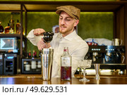 Bearded man in white shirt and cap squeezing lemon juice over shaker with cocktail while working behind counter in pub. Male barkeeper squeezing lemon into cocktail in a pub. Стоковое фото, агентство Ingram Publishing / Фотобанк Лори