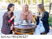 Smiling multiracial female friends eating and drinking coffee while sitting at table on terrace of outdoor cafeteria on summer day. Laughing diverse women in outdoor cafe. Стоковое фото, агентство Ingram Publishing / Фотобанк Лори