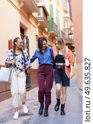 Full length group of diverse female friends in casual clothes speaking happily while passing by urban building. Stylish multiracial ladies walking home together. Стоковое фото, агентство Ingram Publishing / Фотобанк Лори