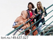 Low angle of joyful young Muslim woman in hijab, laughing while watching photo on smartphone with happy female friends, standing together on metal staircase on sunny day. Diverse cheerful female students smiling while sharing smartphone on staircase. Стоковое фото, агентство Ingram Publishing / Фотобанк Лори