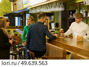 Man ordering beer from barkeeper while standing near happy talking women on weekend day in pub. Clients and bartender in an elegant bar. Стоковое фото, агентство Ingram Publishing / Фотобанк Лори