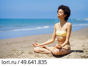 Full body of peaceful slim black female tourist in bikini sitting in Lotus pose on sandy seashore with closed eyes, while meditating during yoga session on sunny summer day. Fit young African American woman in Padmasana meditating on sandy beach. Стоковое фото, агентство Ingram Publishing / Фотобанк Лори