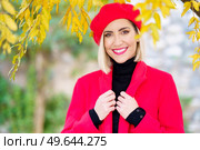 Charismatic young happy female, with blond hair and makeup in elegant red beret and coat smiling and looking at camera standing under tree with yellow leaves in autumn park. Joyful trendy lady smiling at camera in autumn garden. Стоковое фото, агентство Ingram Publishing / Фотобанк Лори