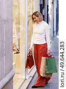 Full body of smiling female customer with purchases in paper shopping bags choosing goods through glass wall of store. Female shopper with bags looking through shop showcase. Стоковое фото, агентство Ingram Publishing / Фотобанк Лори