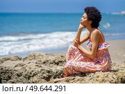 Side black African American female traveler in maxi dress, with curly hair leaning on hand and closing eyes while relaxing on rough rocky shore on blurred background of waving sea. Black woman resting near waving sea. Стоковое фото, агентство Ingram Publishing / Фотобанк Лори
