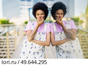 Cheerful African American female touching cheeks while making funny face and looking at camera near building with glass wall in city against blurred background. Positive black woman grimacing near building. Стоковое фото, агентство Ingram Publishing / Фотобанк Лори