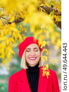 Happy female in bright red beret and coat standing with closed eyes under tree with yellow leaves in park on autumn day against blurred background. Cheerful woman near autumn leaves with red clothes. Стоковое фото, агентство Ingram Publishing / Фотобанк Лори