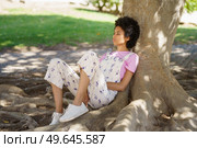 Full body side view of young wistful African American female in casual clothes sitting near thick tree trunk in park. Pensive black woman sitting near tree. Стоковое фото, агентство Ingram Publishing / Фотобанк Лори