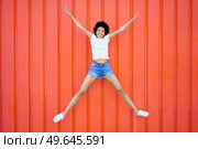 Full body of optimistic African American female in casual clothes looking at camera while star jumping on red background in city. Joyful black woman jumping near wall. Стоковое фото, агентство Ingram Publishing / Фотобанк Лори