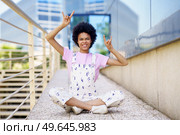 Full body of carefree African American female in casual outfit looking at camera with rock and roll gestures while sitting on street. Rebellious black woman showing horn signs. Стоковое фото, агентство Ingram Publishing / Фотобанк Лори