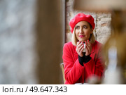 Through blurred column and lamp of charming female in red clothes looking away while sitting with cup of hot drink in modern cafeteria. Dreamy woman with coffee in cafe. Стоковое фото, агентство Ingram Publishing / Фотобанк Лори