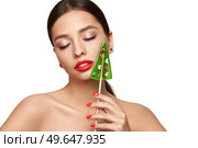 Beauty model girl eating Christmas tree shape lollipop.  Beautiful young woman with clean fresh skin. Girl beauty face care. Facial treatment. Cosmetology, beauty and spa. Red nails manicure. Стоковое фото, агентство Ingram Publishing / Фотобанк Лори