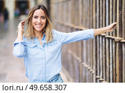 Happy adult female in stylish blue shirt looking at camera with smile and touching hair while grasping metal fence on blurred background of city street. Cheerful woman touching hair near fence. Стоковое фото, агентство Ingram Publishing / Фотобанк Лори