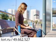 Female manager in stylish dress and sunglasses sitting on bench near suitcase and thermos and browsing netbook on station during business trip in city. Businesswoman using laptop on bench. Стоковое фото, агентство Ingram Publishing / Фотобанк Лори