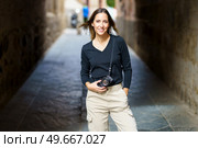 Self assured young female photographer with camera in casual clothes standing on narrow street with hand in pocket and looking at camera during vacation. Smiling woman with photo camera looking at camera on narrow street. Стоковое фото, агентство Ingram Publishing / Фотобанк Лори