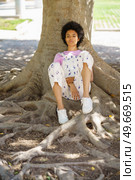 Full body of dreamy African American female looking away while sitting near thick tree trunk with roots in park on summer day. Pensive black woman sitting near tree. Стоковое фото, агентство Ingram Publishing / Фотобанк Лори