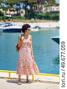 Full body black woman in long dress and sunglasses carrying bag on shoulder, and admiring sea while standing on pier near boats on summer weekend day in town port. Ethnic traveler standing on quay in harbor. Стоковое фото, агентство Ingram Publishing / Фотобанк Лори