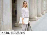 Serious self assured adult woman with blond hair in gray outfit standing on street near old building and holding laptop and handbag while looking away. Confident businesswoman with laptop on street. Стоковое фото, агентство Ingram Publishing / Фотобанк Лори