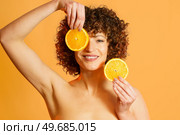 Happy woman with bare shoulders and curly hair, covering eye with slice of citrus and looking at camera with smile during skin care routine against orange background. Cheerful female model with slices of orange. Стоковое фото, агентство Ingram Publishing / Фотобанк Лори