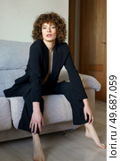 Full body of adult female with dark curly hair wearing black suit sitting on gray sofa leaning forward and looking at camera. Gorgeous woman in suit with bare chest sitting on couch. Стоковое фото, агентство Ingram Publishing / Фотобанк Лори