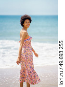 Side view of carefree woman in stylish dress standing on sandy beach washed by rippling sea water and looking at camera. Cheerful ethnic woman enjoying bright summer day at seaside. Стоковое фото, агентство Ingram Publishing / Фотобанк Лори